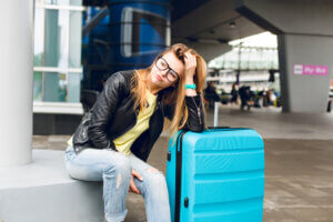 Woman sitting outside an airport with luggage, looking frustrated while waiting for a transfer that has not arrived