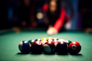 Racked balls in focus with a blurred player preparing to break behind them on a green baize table in low ambient light.