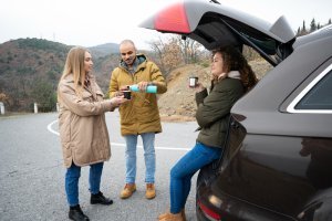 Three people in winter coats sharing a flask of coffee beside an open car boot on a quiet country road, with rolling hills and bare-branched trees in the background, on a relaxed bank holiday weekend trip.