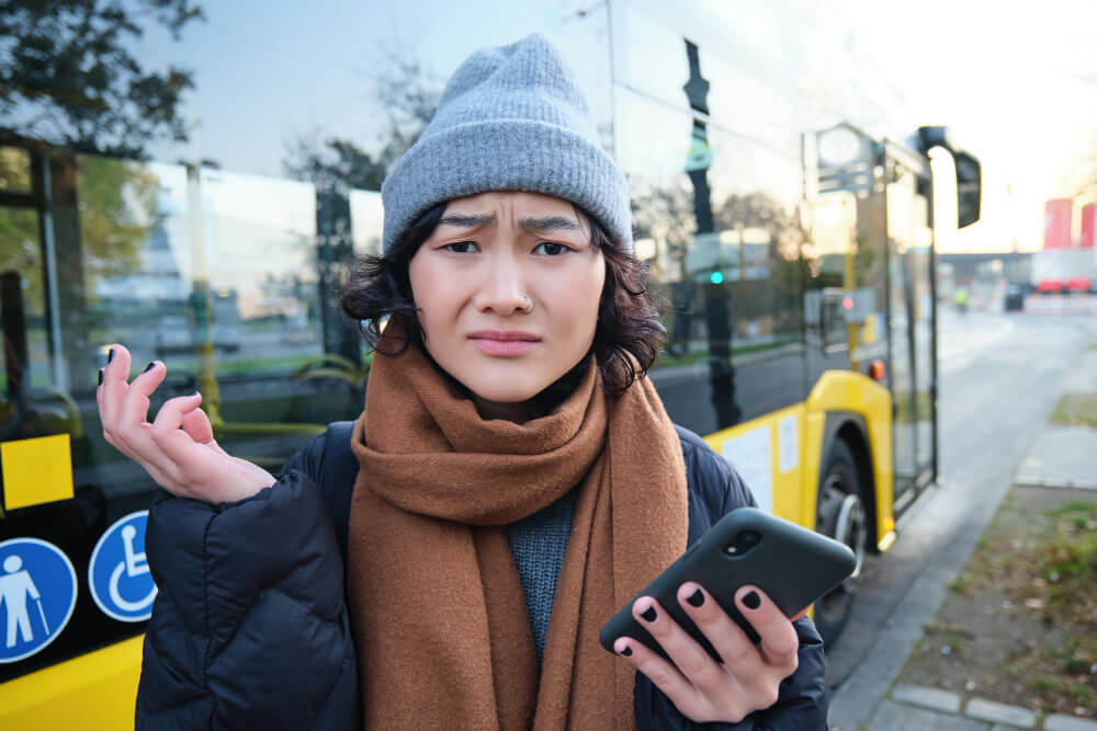 Frustrated traveller holding a smartphone beside a bus, unable to locate the correct address from navigation directions