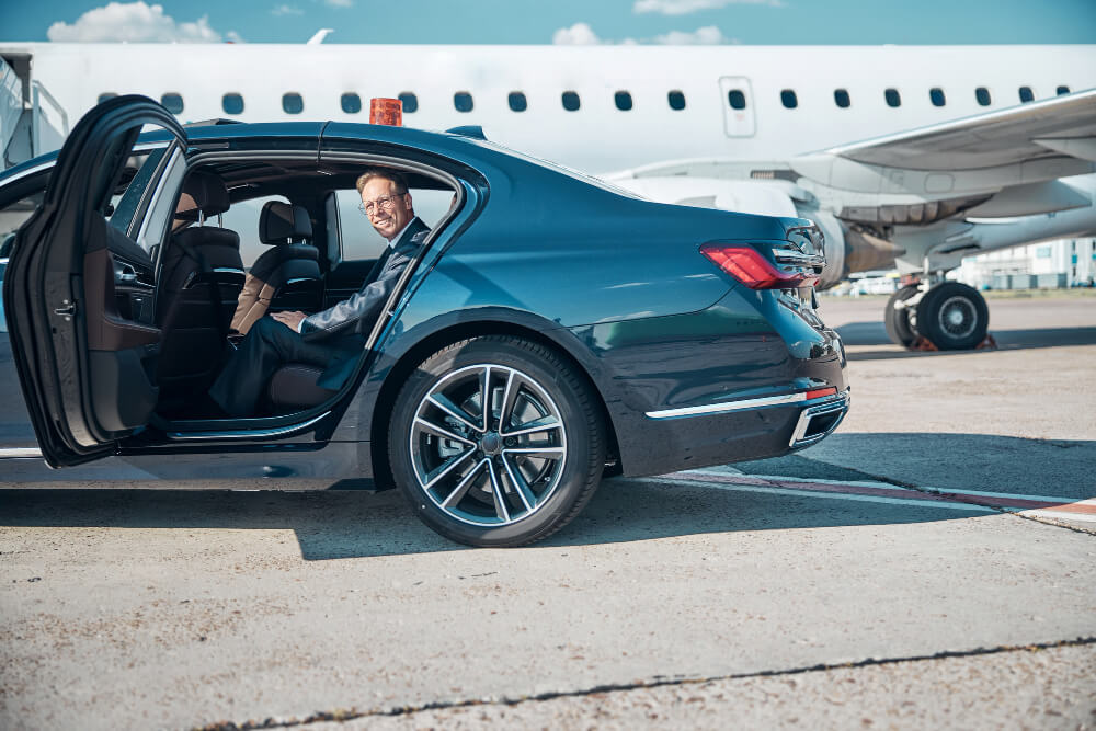 Business executive stepping out of dark executive saloon beside a private jet on the apron at a London private aviation airport