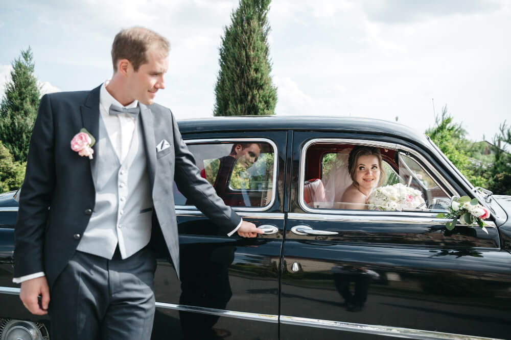 Groom opening the door of a black car for a bride holding a bouquet, arriving at a wedding venue