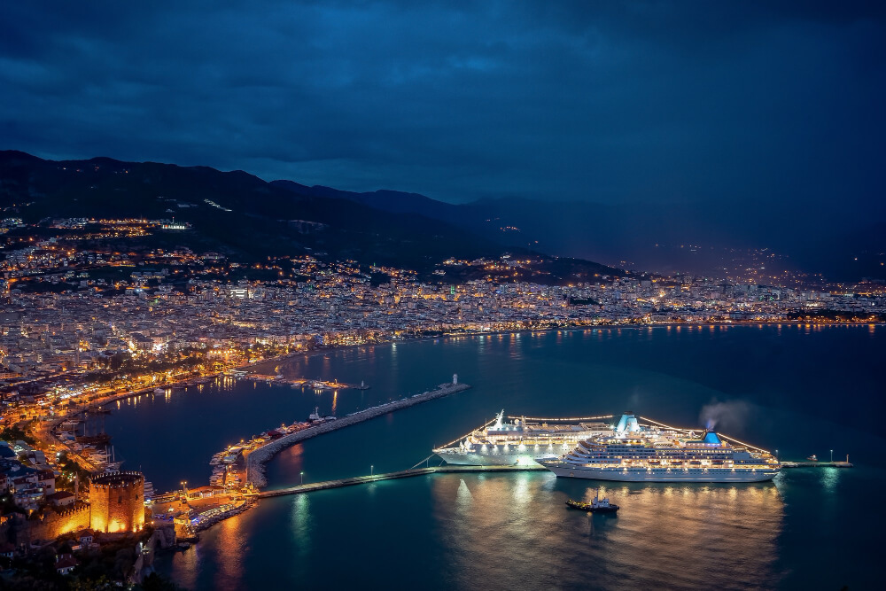 Aerial night view of a cruise ship illuminated at a coastal port terminal with city lights stretching to the hills beyond