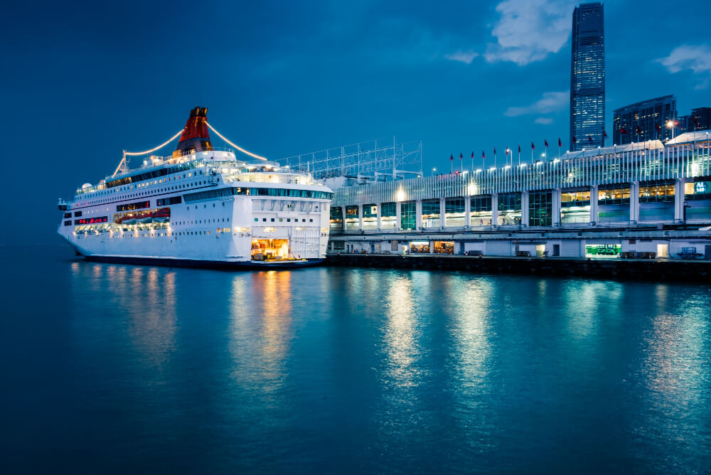 Cruise ship moored at an illuminated terminal at night, lights reflected in calm harbour water the evening before departure