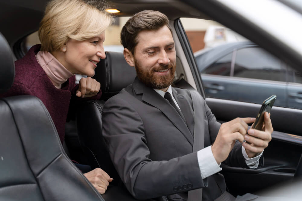 Two passengers in the rear seat of a private transfer vehicle in London, relaxed and looking at a smartphone during their journey
