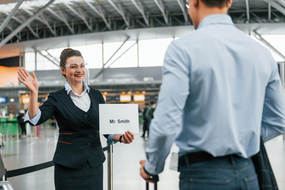 A driver in an airport arrivals hall holding a name board as passengers walk out of customs