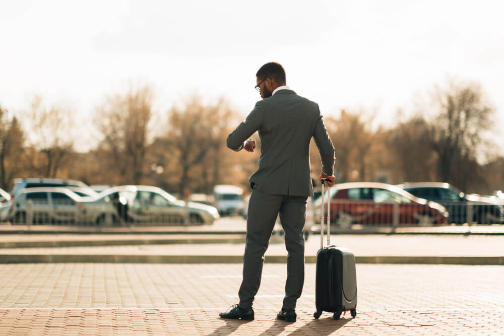 Business traveller in a suit with a suitcase checking his watch in an airport car park, waiting for a pre-booked transfer
