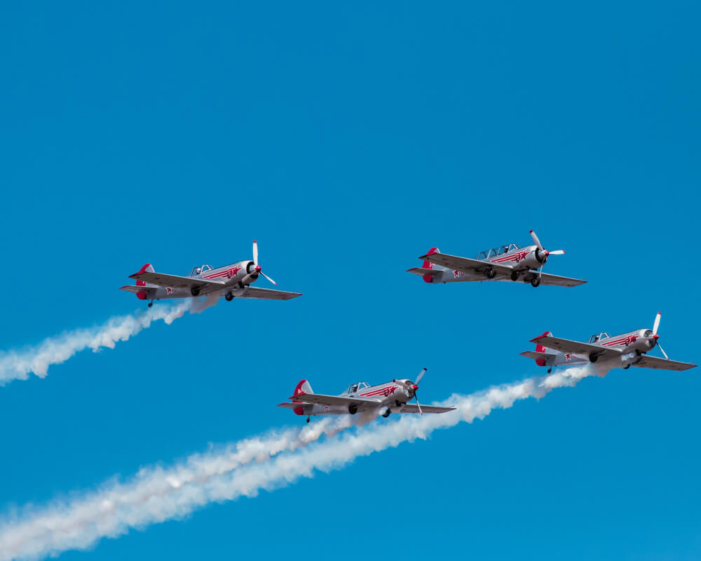 Four red and silver propeller aircraft flying in diamond formation with white smoke trails at Farnborough Airshow flying display